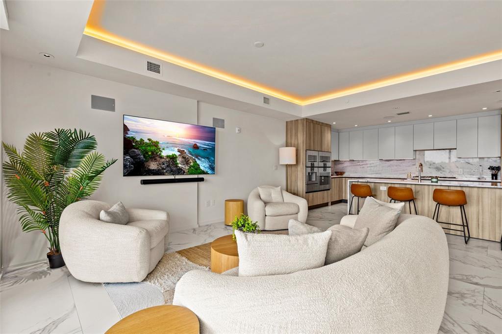 A modern open-concept living room features a curved sofa, armchairs, and a kitchen with stools — 1020 SUNSET POINT ROAD UNIT 310, CLEARWATER, FL