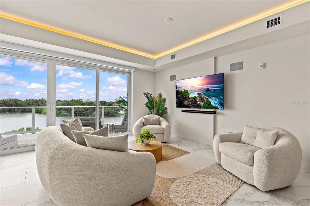 A modern living room features white curved furniture, a wall-mounted television, and floor-to-ceiling windows — 1020 SUNSET POINT ROAD UNIT 310, CLEARWATER, FL