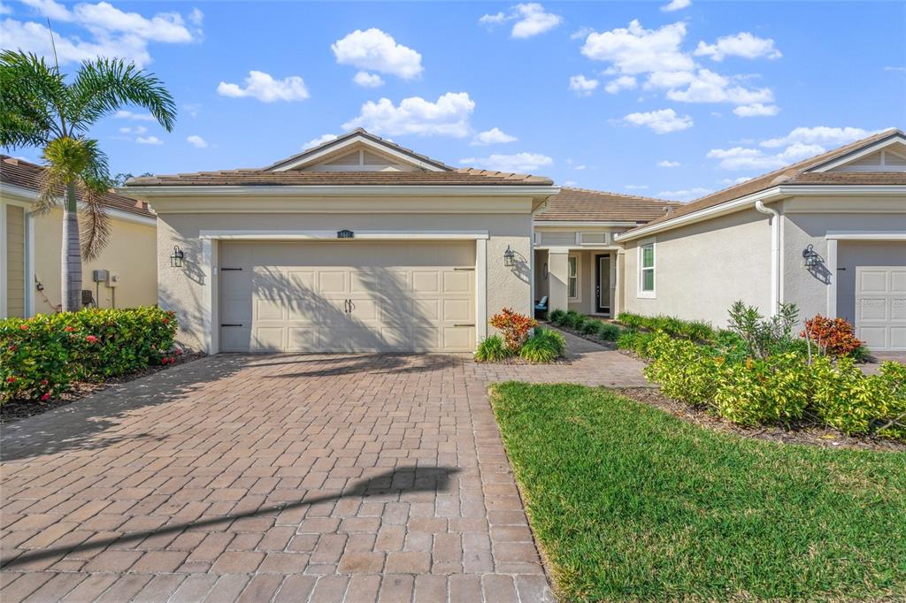 A beige single-story house features a paver driveway, a two-car garage, and landscaped front gardens — 8681 RAIN SONG ROAD, SARASOTA, FL
