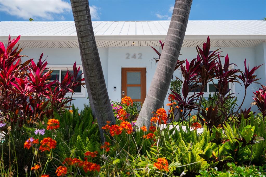 A light blue house exterior with a brown door, palm trees, and vibrant tropical landscaping — 242 GLADIOLUS STREET, ANNA MARIA, FL