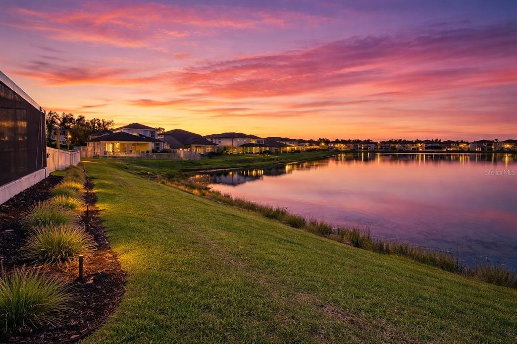 A grassy lawn slopes down to a lake at sunset with houses in the background — 3103 MARINE GRASS DRIVE, WIMAUMA, FL