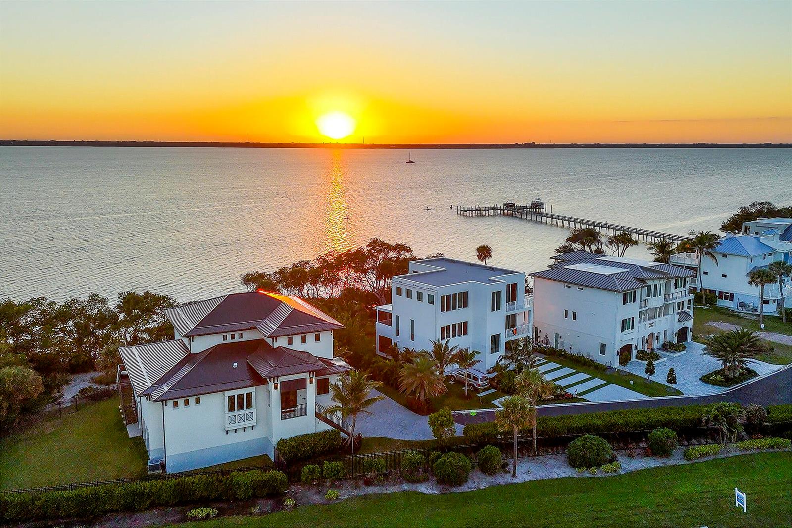 An aerial view of three waterfront houses at sunset with a long wooden pier nearby — 7683 Pelican Pointe Drive, Jensen Beach FL 34957, JENSEN BEACH, FL
