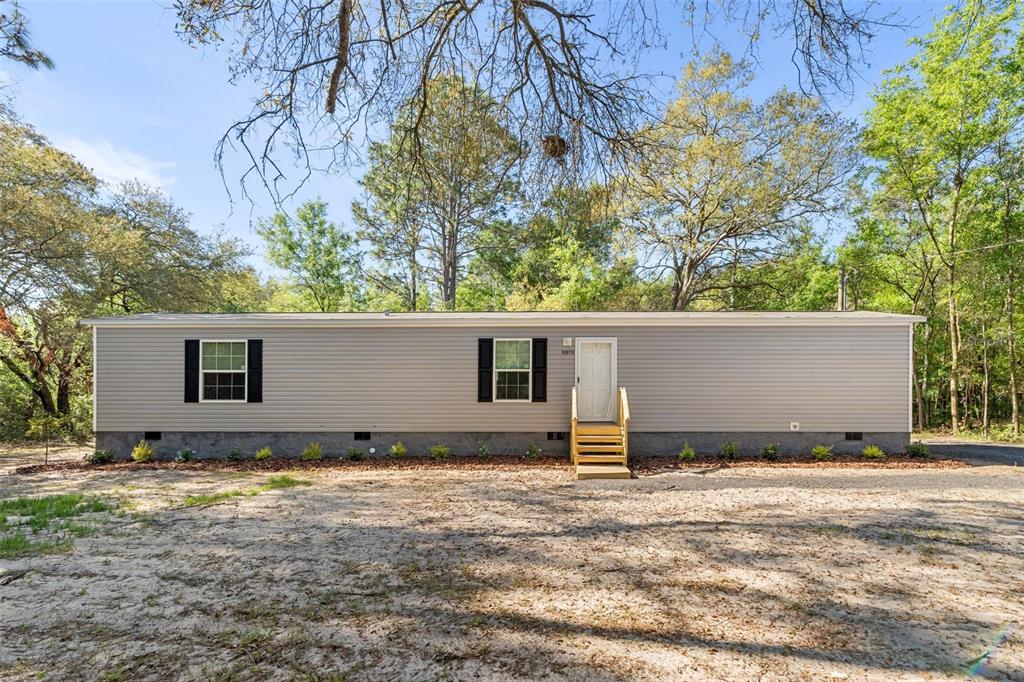A single-story manufactured home with gray siding and black shutters sits on a sandy lot — 10970 NE 71ST STREET, BRONSON, FL