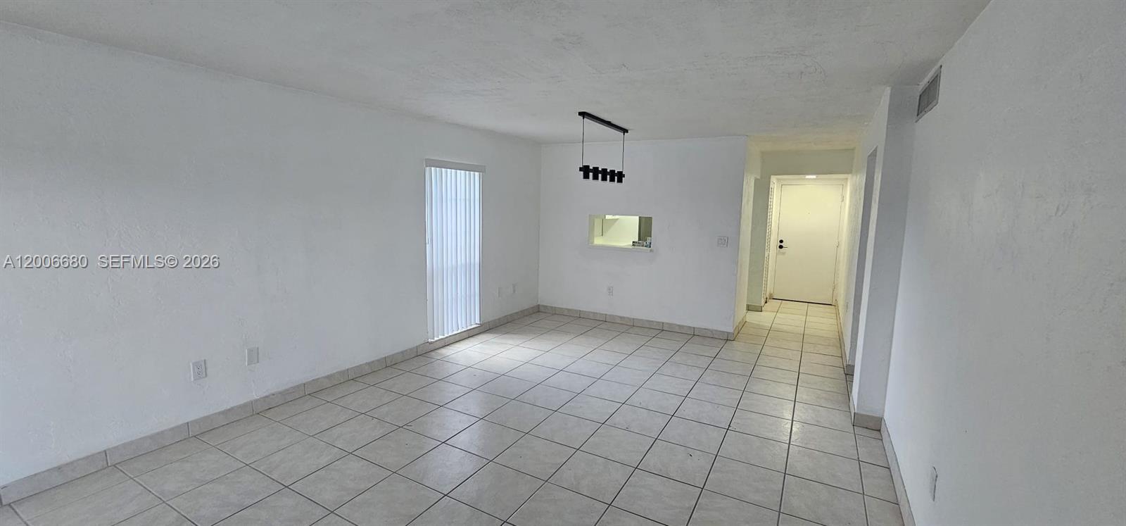 An empty room features white walls, light-colored tile flooring, a vertical blind window, and a chandelier — 6884 N Kendall Dr # C404, Pinecrest FL 33156, PINECREST, FL