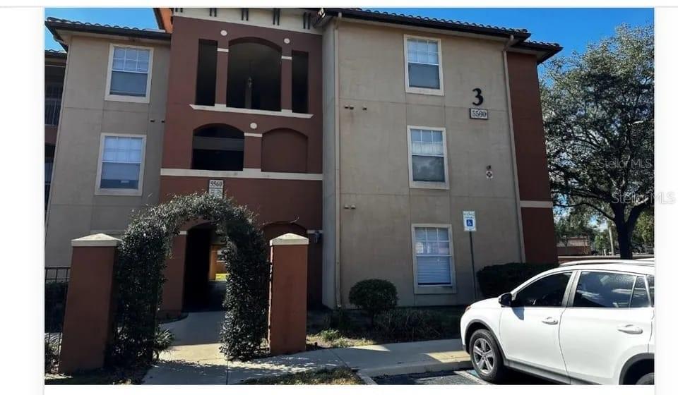A three-story apartment building exterior with a gated walkway and a parked white car — 5560 METROWEST BOULEVARD UNIT 3-207, ORLANDO, FL