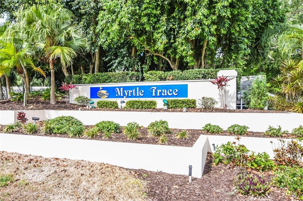 A white tiered retaining wall features a blue sign reading Myrtle Trace amidst lush landscaping — 339 PEMBROKE LANE S UNIT 228, VENICE, FL