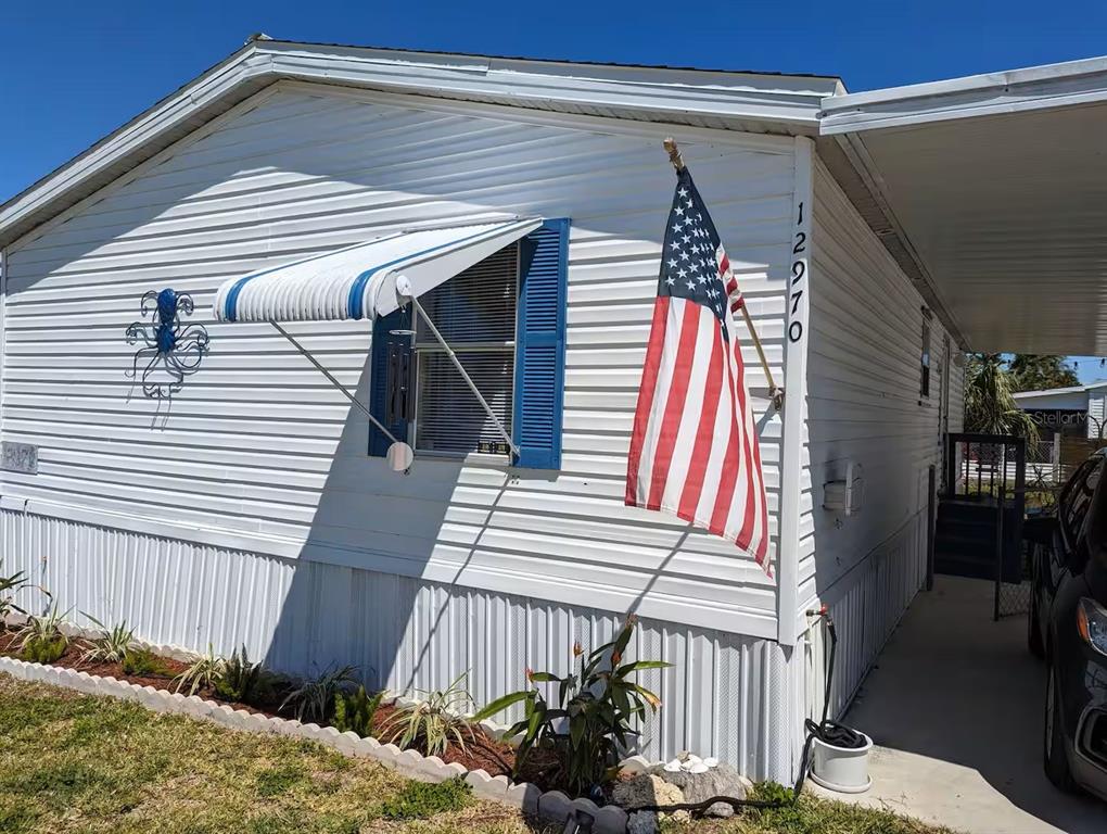 A white mobile home exterior features a blue-shuttered window, an American flag, and landscaping — 12970 SPENCER STREET, FORT MYERS, FL