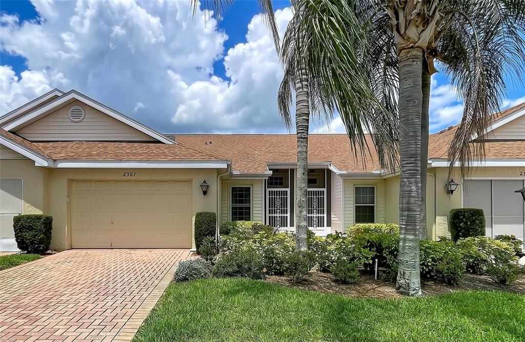 A single-story house with a tan garage, brick paver driveway, and palm trees in front — 2307 NEW ORCHARD COURT, SUN CITY CENTER, FL