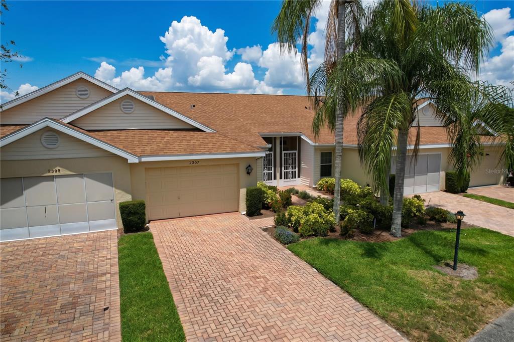 An exterior view of a tan, single-story duplex with a brick driveway and palm tree — 2307 NEW ORCHARD COURT, SUN CITY CENTER, FL