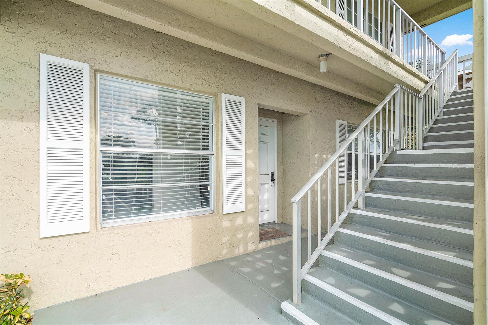 An exterior view of a tan stucco building with a white door, window, and staircase — 1021 Flame Vine Avenue # 104, Delray Beach FL 33445, DELRAY BEACH, FL