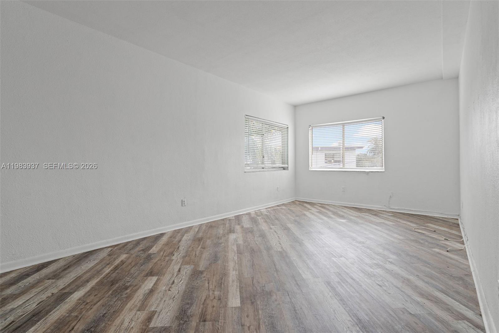 An empty room features white walls, wood-look laminate flooring, and two windows with blinds — 1185 Marseille Dr # 202, Miami Beach FL 33141, MIAMI BEACH, FL