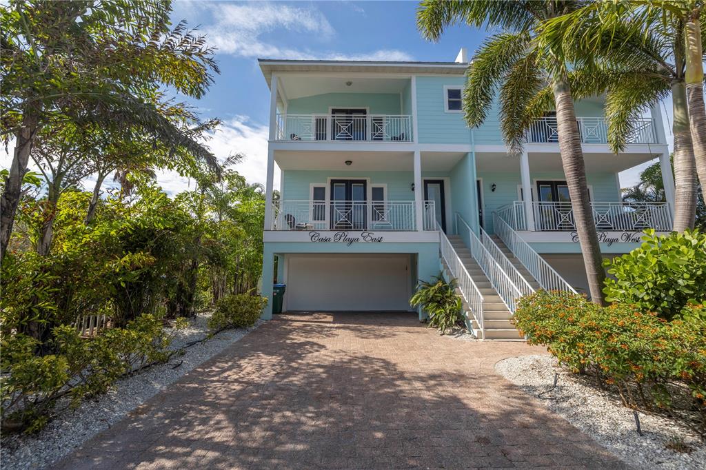 A light blue duplex house with elevated balconies and a paved driveway sits among palm trees — 105 4TH STREET S, BRADENTON BEACH, FL