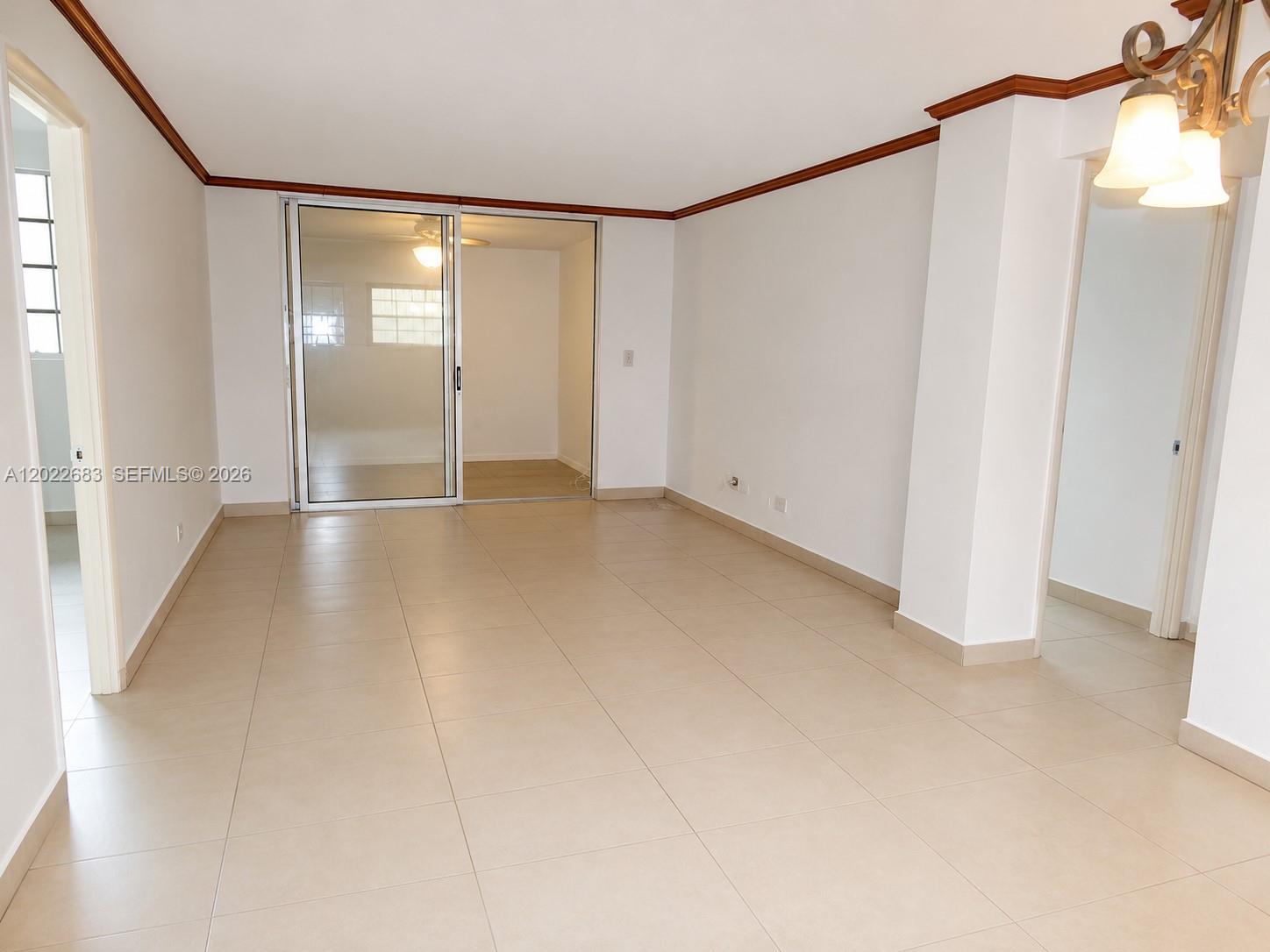 An empty room features light-colored tile flooring, white walls, crown molding, and a sliding glass door — 447 NE 195th St # 117, Miami FL 33179, MIAMI, FL