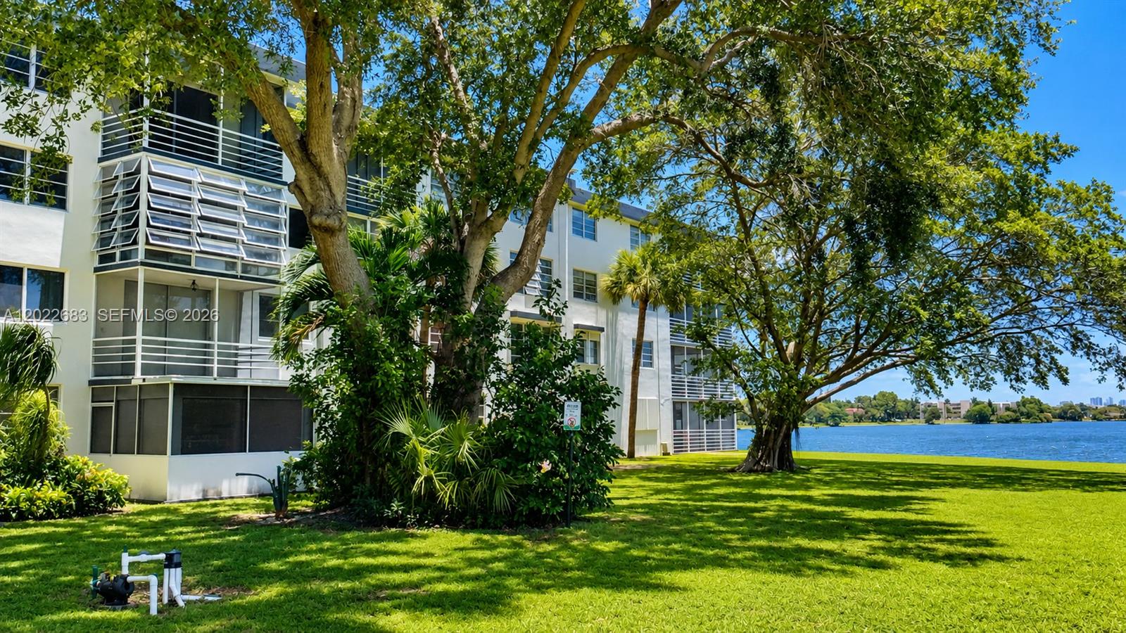 A multi-story apartment building sits beside a lake surrounded by large trees and grass — 447 NE 195th St # 117, Miami FL 33179, MIAMI, FL