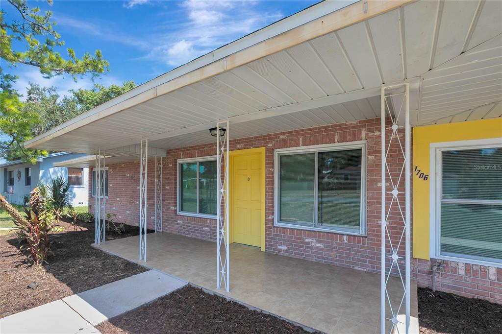A brick house exterior features a covered porch with white metal columns and a yellow door — 1706 SHORE ACRES DRIVE, LAKELAND, FL