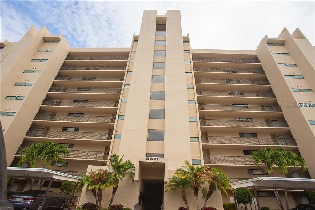 A low-angle view of a multi-story beige apartment building with balconies and palm trees — 2621 COVE CAY DRIVE UNIT 105, CLEARWATER, FL