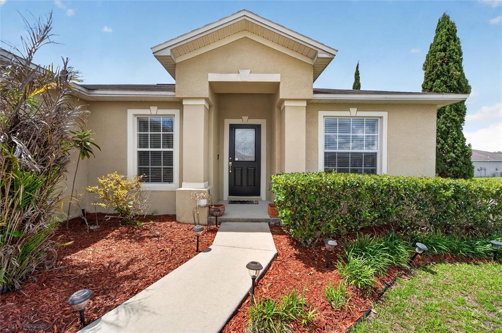 The exterior of a single-story house features a tan stucco facade, a dark front door, and landscaping — 603 POINTER LANE, LAKELAND, FL