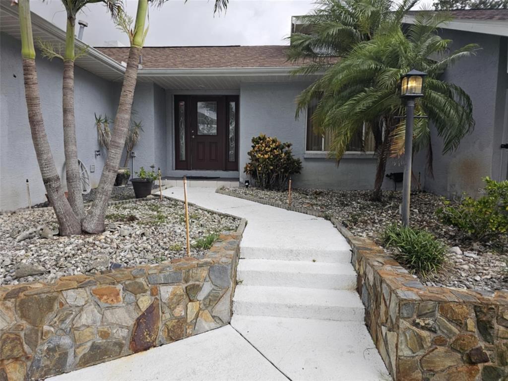 A stone-walled walkway with steps leads to the front entrance of a gray house — 2210 BAYOU DRIVE, HOLIDAY, FL