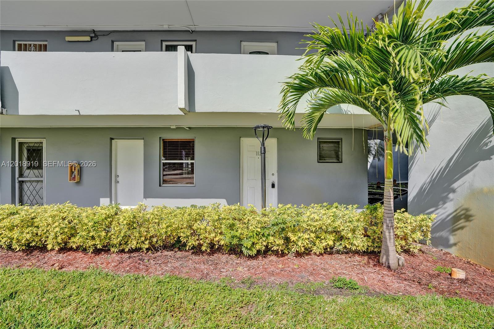 The exterior of a two-story apartment building features a palm tree and a landscaped hedge — 1601 NE 191 ST # B112, Miami FL 33179, MIAMI, FL
