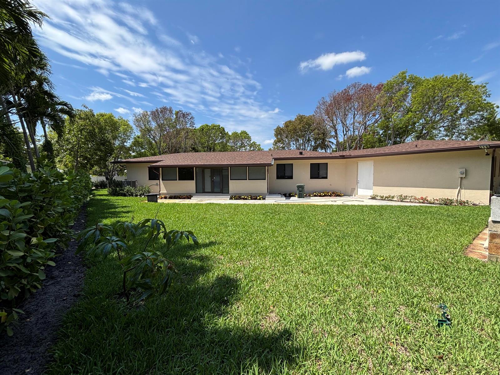 A backyard view of a single-story house with a lawn, patio, and surrounding trees — 135 E Ilex Drive, Lake Park FL 33403, LAKE PARK, FL