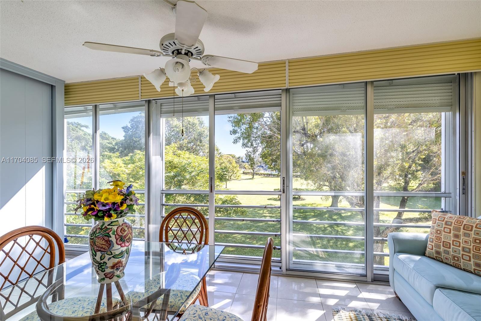 A dining area features a glass table, wicker chairs, and large sliding windows overlooking trees — 3029 Berkshire B # 3029, Deerfield Beach FL 33442, DEERFIELD BEACH, FL