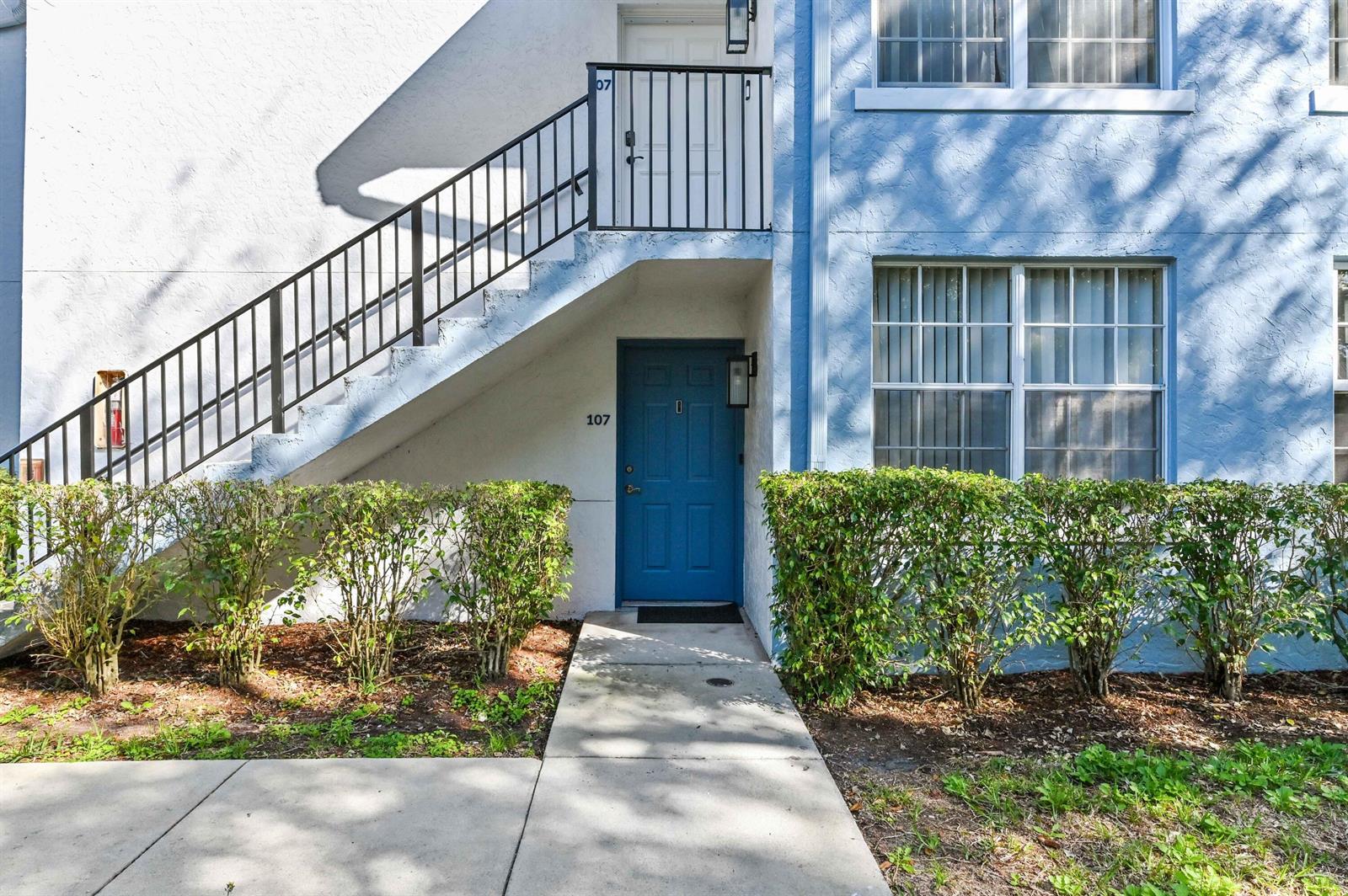 A blue exterior apartment building features a ground-floor entrance, a staircase, and landscaped bushes — 3237 Clint Moore Road # 107, Boca Raton FL 33496, BOCA RATON, FL