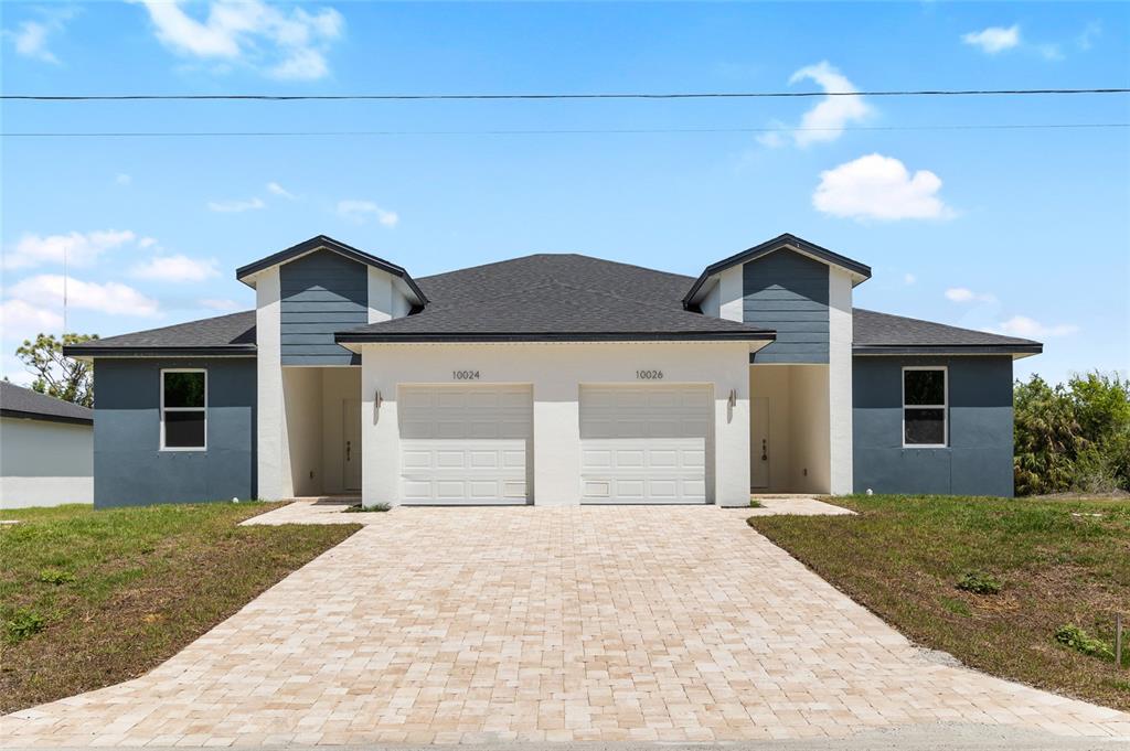 A duplex features a shared paver driveway, white garage doors, and dark blue exterior siding — 10026 COCOA BEACH STREET, PORT CHARLOTTE, FL