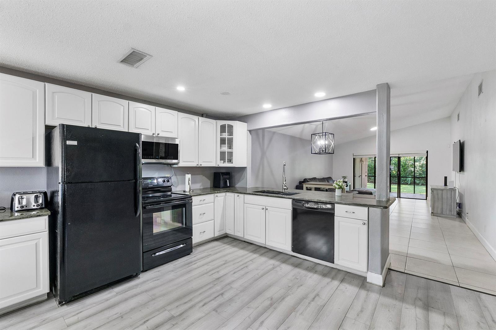 A kitchen with white cabinets, black appliances, and light wood-look flooring opens to a living area — 4149 Palm Forest Drive S, Delray Beach FL 33445, DELRAY BEACH, FL