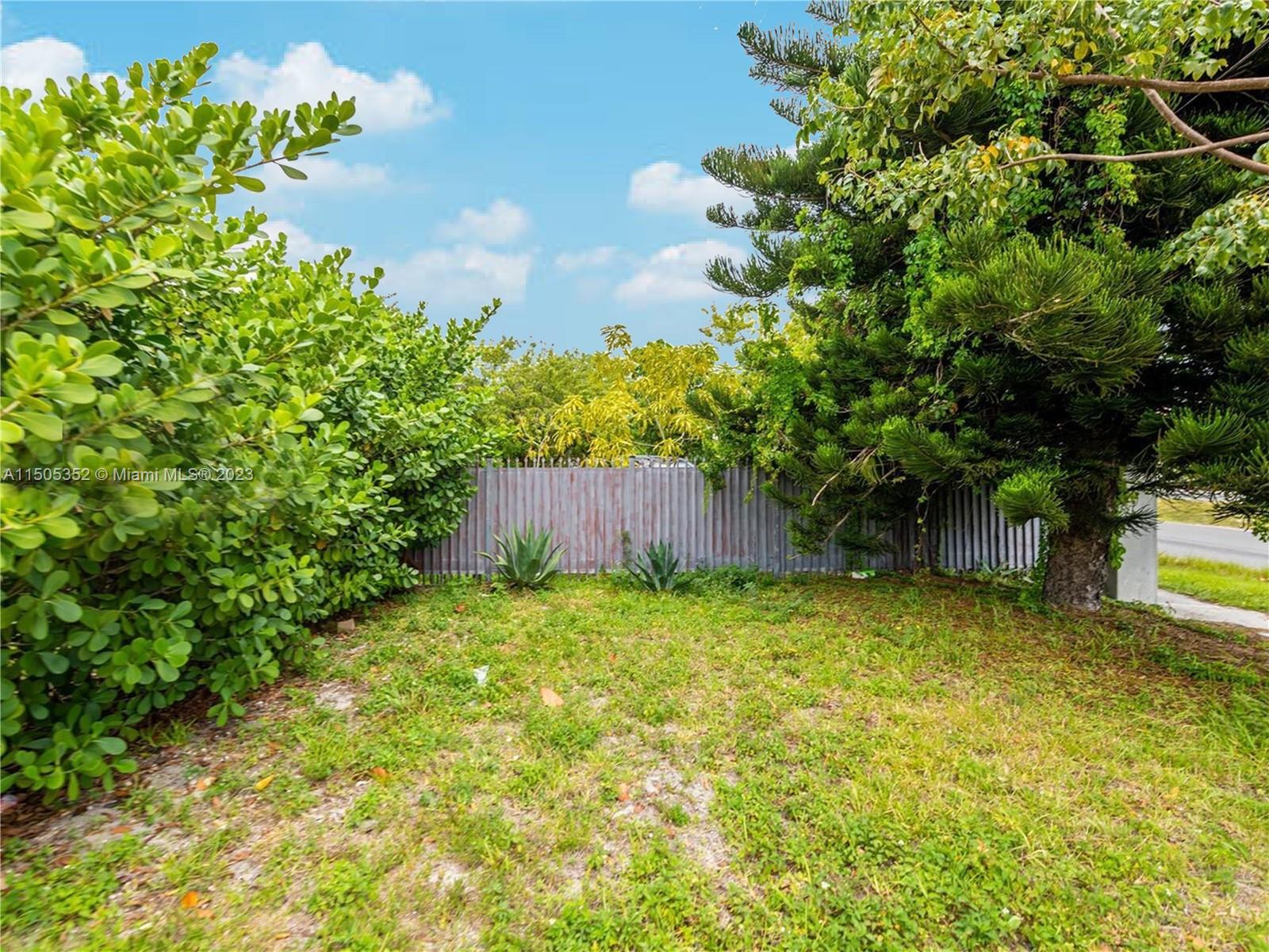 A grassy backyard features a corrugated metal fence surrounded by trees and shrubs — 400 1st St, Dania Beach FL 33004, DANIA BEACH, FL