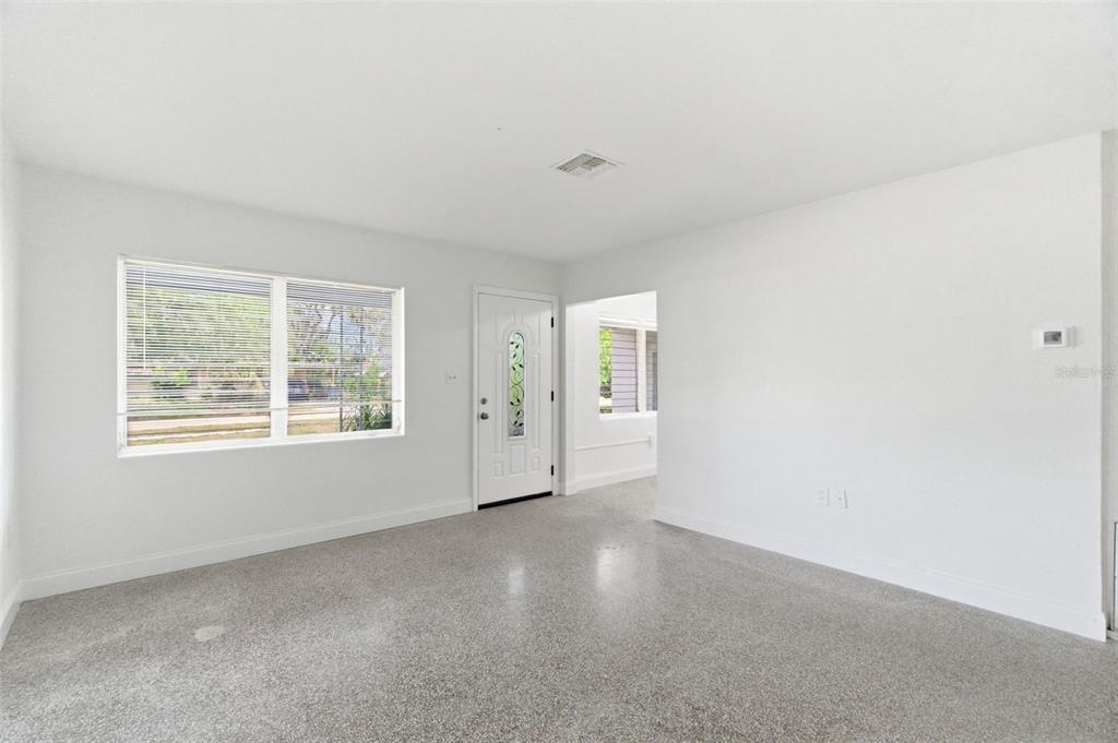 A bright, empty room features white walls, a front door, and speckled gray flooring — 7204 ASTOR DRIVE, NEW PORT RICHEY, FL