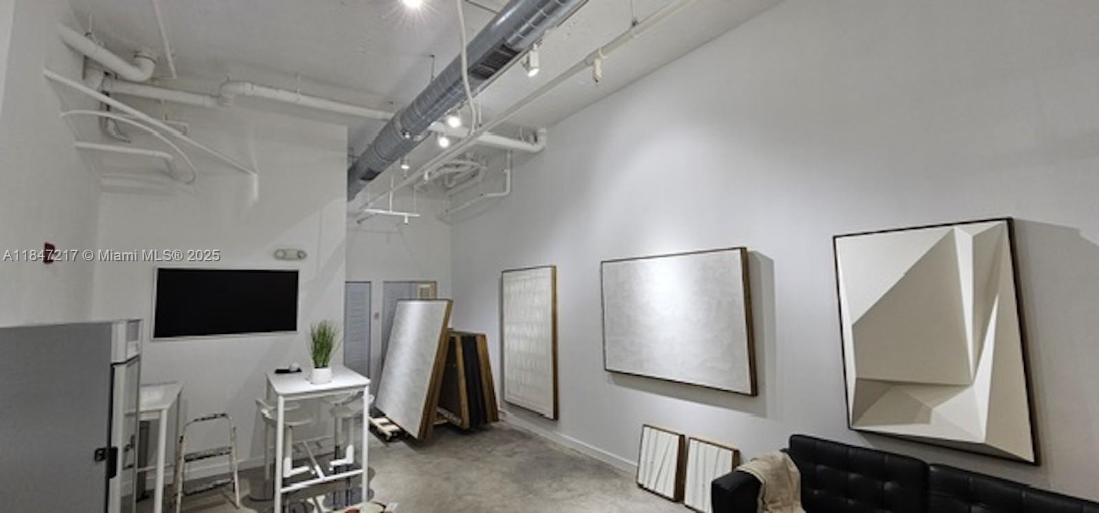 An industrial-style room features white walls, exposed ceiling pipes, concrete floors, and framed artwork — 136 NW 26th St, Miami FL 33127, MIAMI, FL