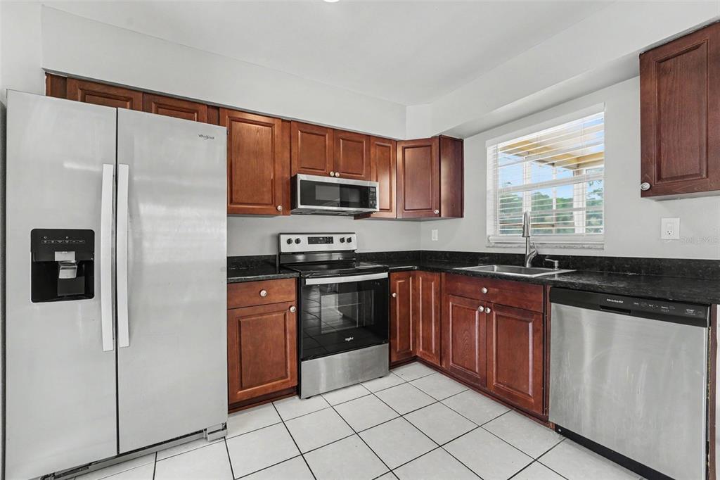 A kitchen with wooden cabinets, stainless steel appliances, black countertops, and white tile flooring — 8332 FISHHAWK AVENUE, NEW PORT RICHEY, FL