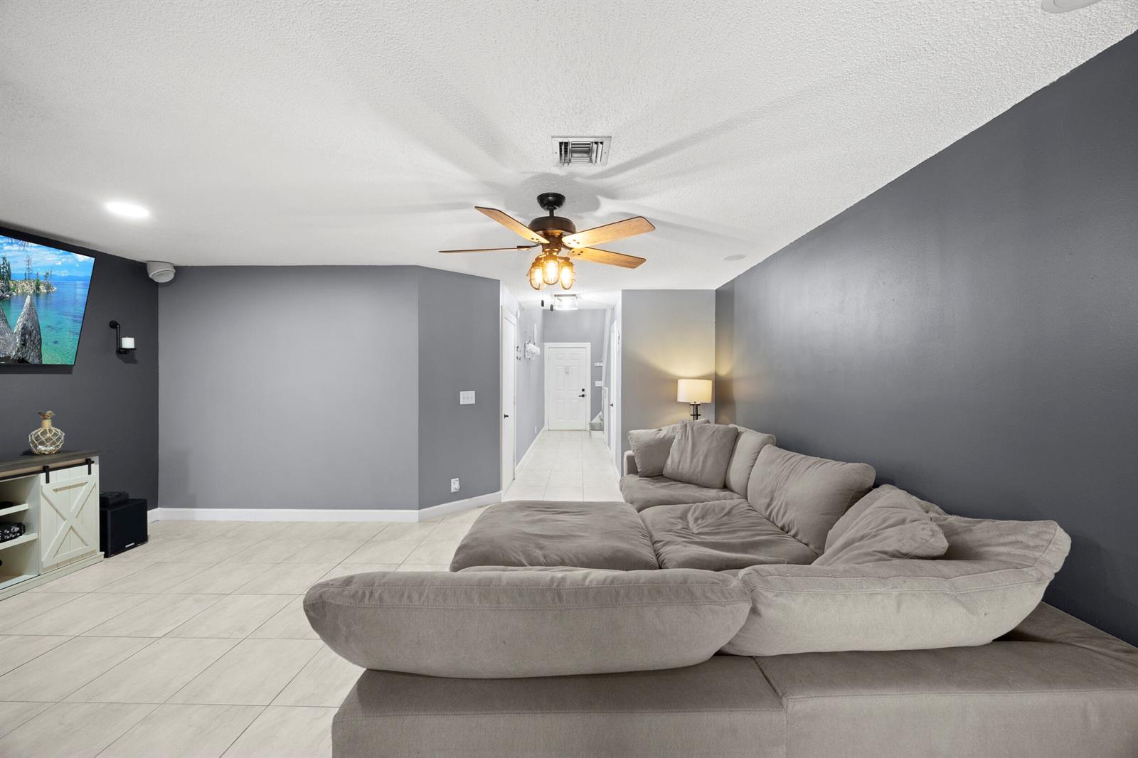 A living room features a grey sectional sofa, a wall-mounted television, and tile flooring — 1928 Discovery Circle E, Deerfield Beach FL 33442, DEERFIELD BEACH, FL