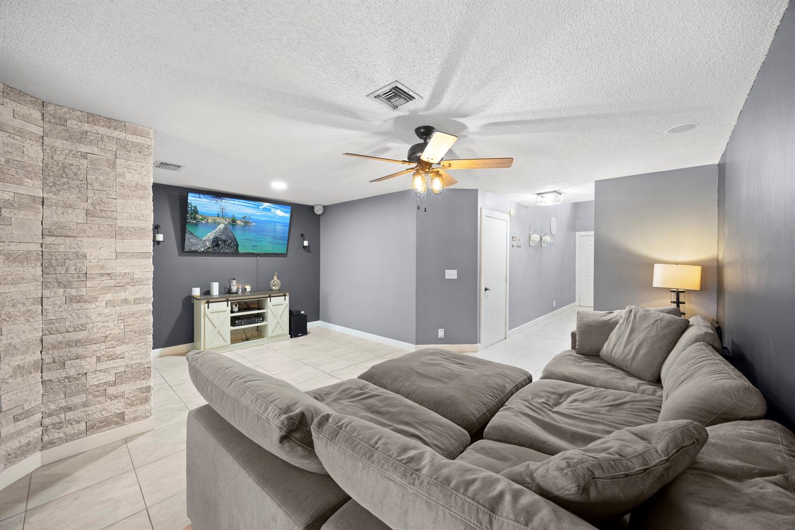 A living room features a gray sectional sofa, a wall-mounted television, and a stone accent wall — 1928 Discovery Circle E, Deerfield Beach FL 33442, DEERFIELD BEACH, FL