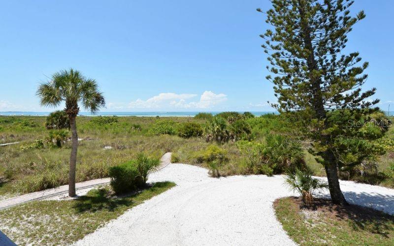 A gravel path leads through coastal vegetation toward a beach under a clear blue sky — 460 BEACH ROAD UNIT A1, SARASOTA, FL