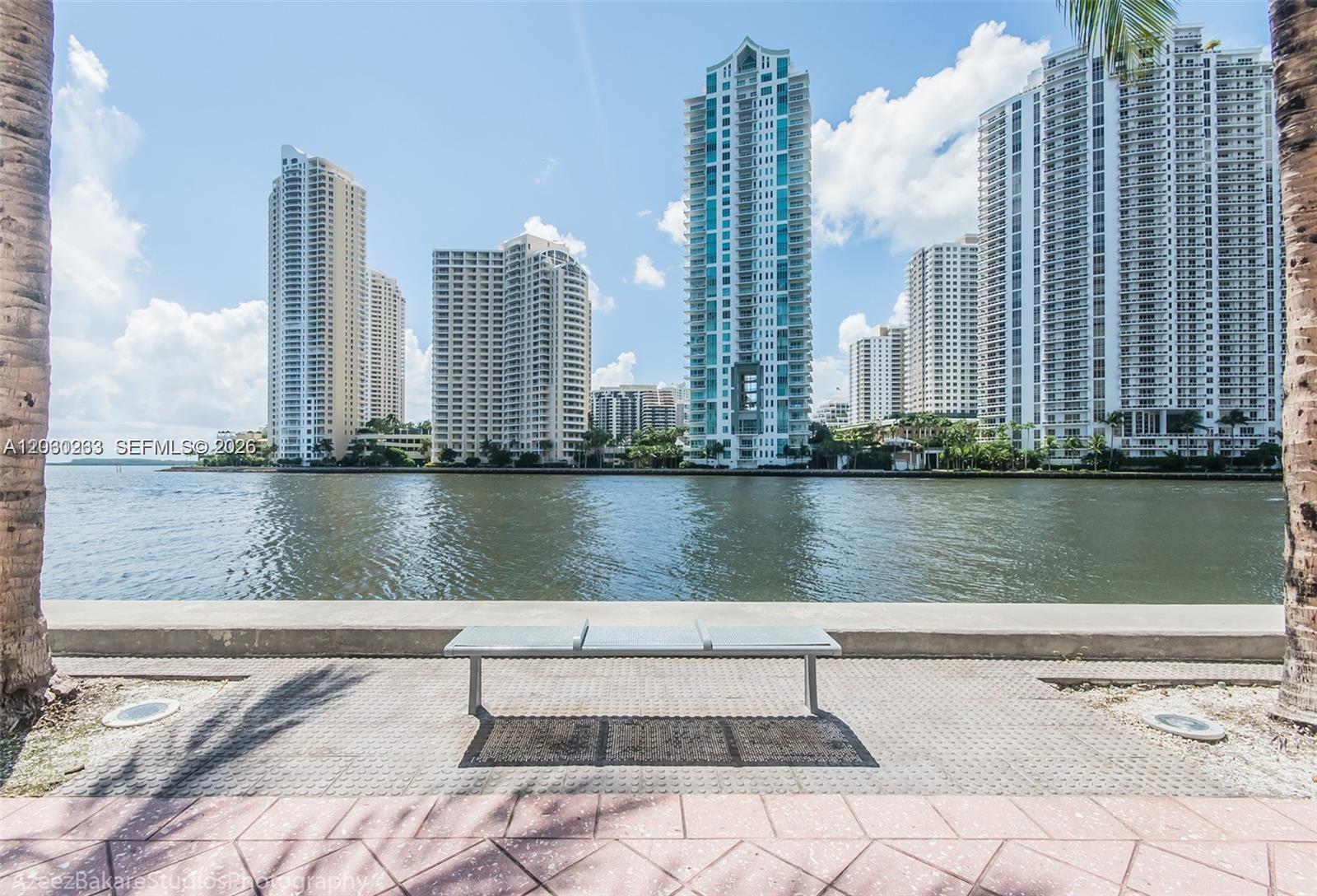 A metal bench sits on a paved waterfront walkway facing a skyline of tall buildings — 335 S BISCAYNE BLVD # 2908, Miami FL 33131, MIAMI, FL