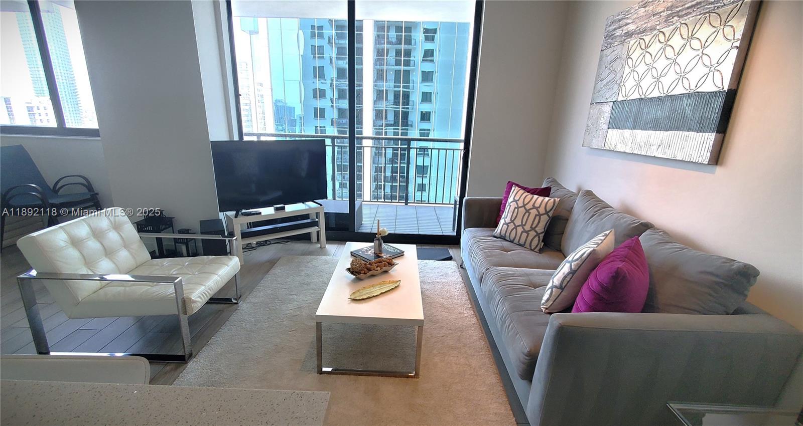 A modern living room features a grey sofa, white accent chair, and floor-to-ceiling balcony windows — 999 SW 1st Ave # 2115, Miami FL 33130, MIAMI, FL