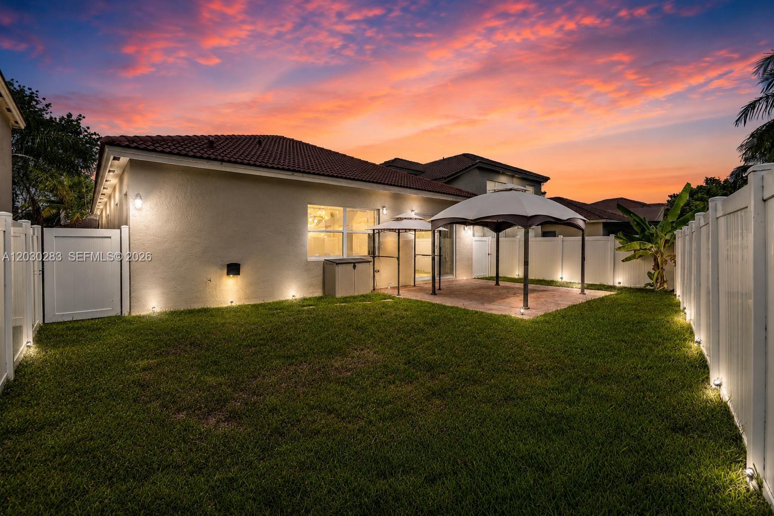 A backyard features a paved patio with a gazebo, lawn, and white vinyl fencing at sunset — 1785 NE 37th Pl, Homestead FL 33033, HOMESTEAD, FL