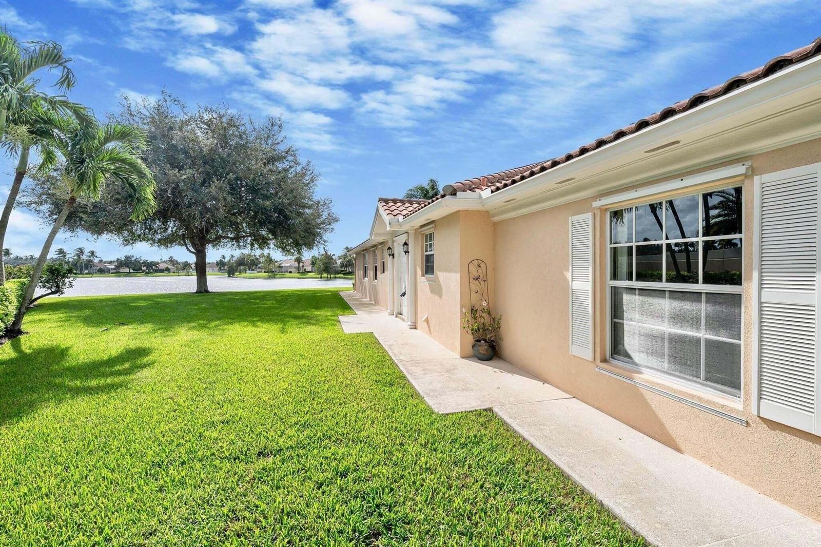 A beige house exterior with a tiled roof sits beside a lawn and a lake — 8270 Eleuthera Lane, Wellington FL 33414, WELLINGTON, FL