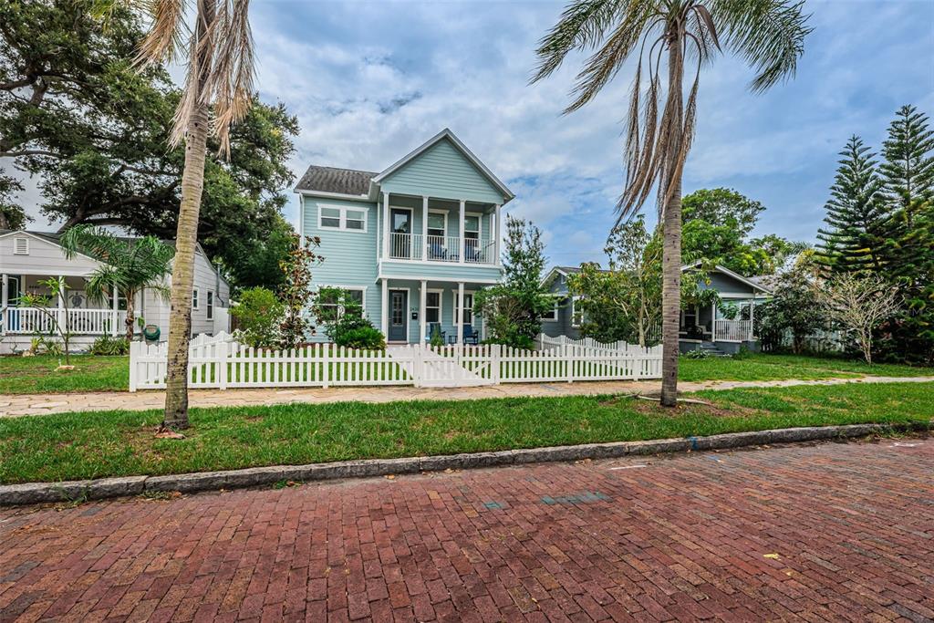 A light blue two-story house with a white picket fence sits behind a brick street — 2430 6TH AVENUE N, ST PETERSBURG, FL