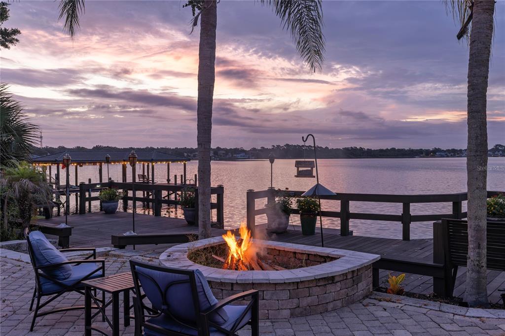 A stone fire pit with chairs sits on a patio overlooking a lake and dock — 2201 COVE COURT, LONGWOOD, FL