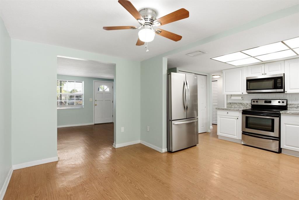 A kitchen with stainless steel appliances, white cabinets, and light wood flooring opens to a room — 7631 HOLLYRIDGE DRIVE, NEW PORT RICHEY, FL