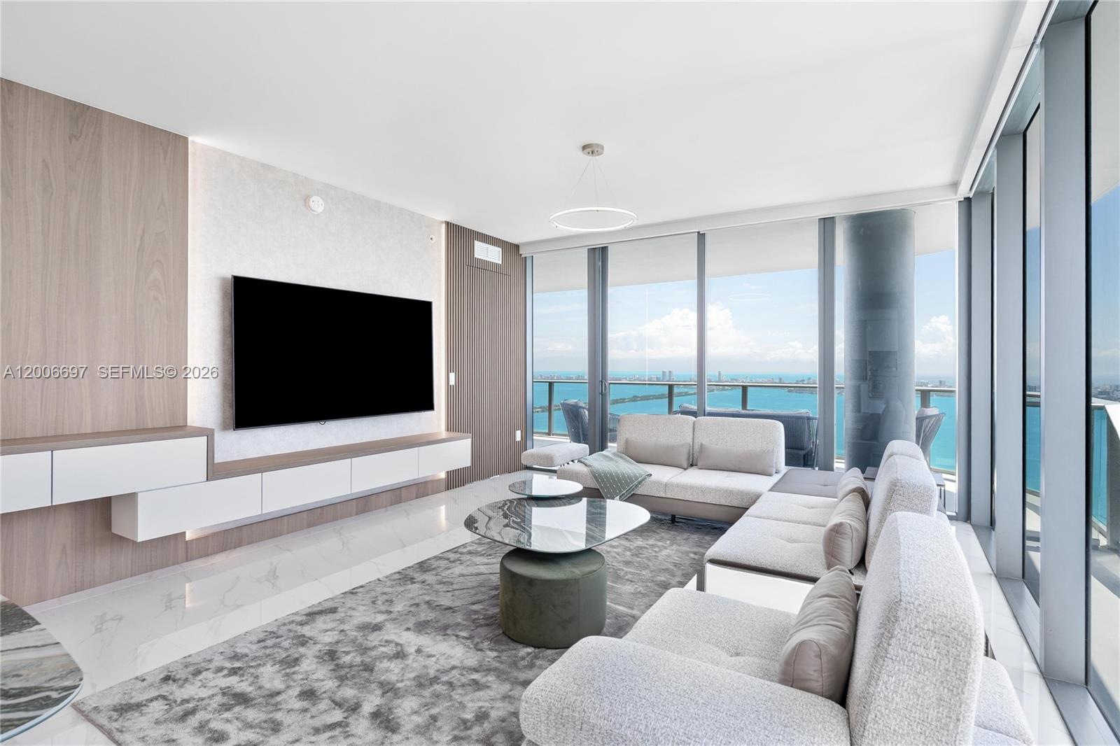 A modern living room features a wall-mounted television, white cabinetry, and floor-to-ceiling windows overlooking water — 700 NE 24th ST # 5407, Miami FL 33137, MIAMI, FL