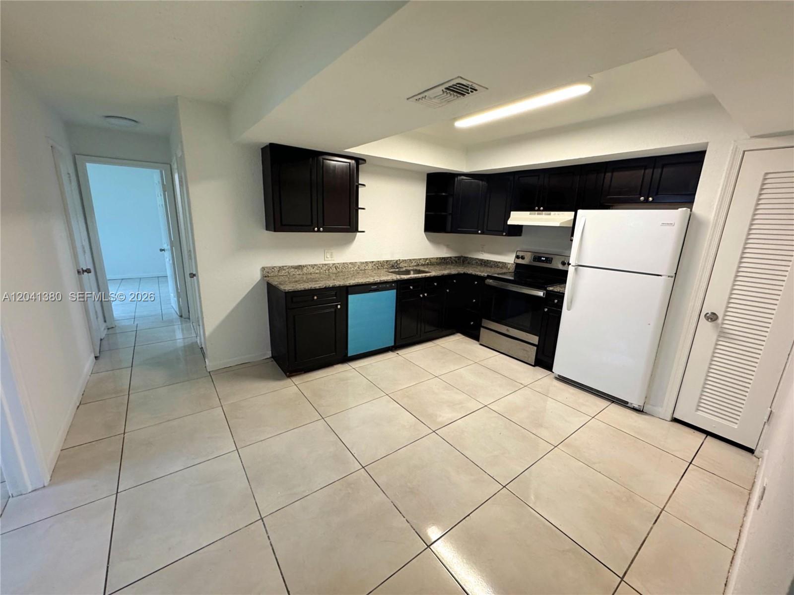 A kitchen features dark cabinets, granite countertops, a white refrigerator, and light-colored tile flooring — NORTH LAUDERDALE, FL
