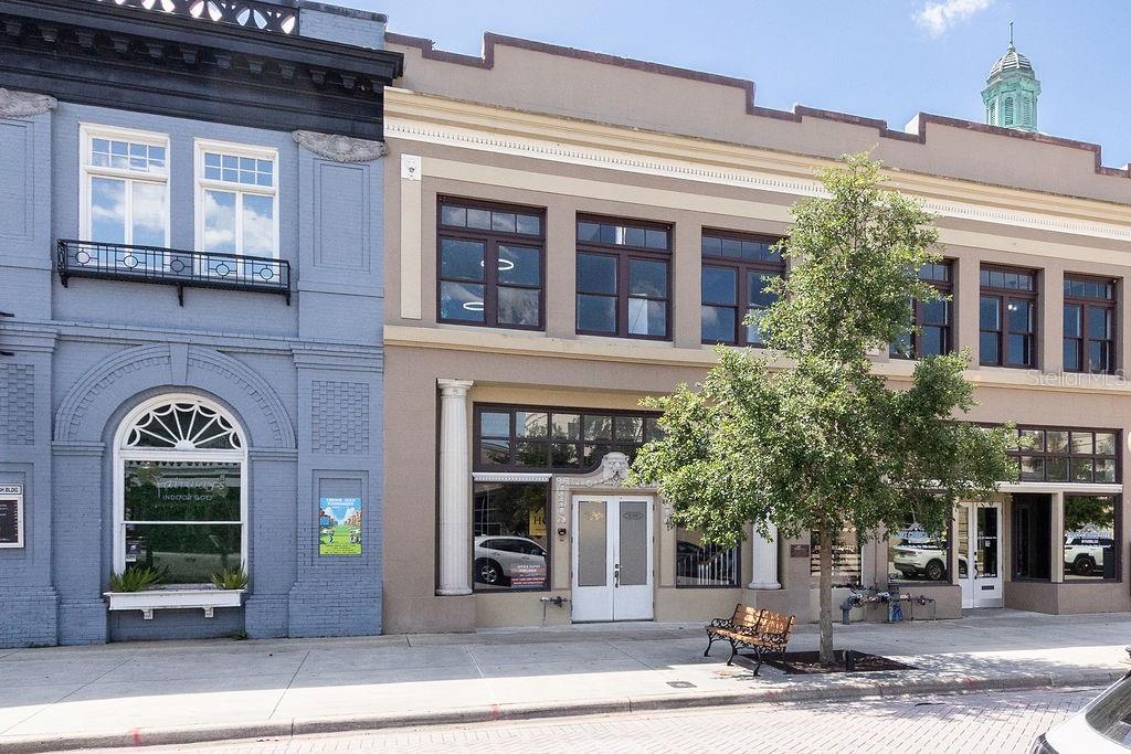 The exterior of two adjacent historic brick buildings features large windows and a sidewalk bench — 112 W INDIANA AVENUE UNIT 206, DELAND, FL