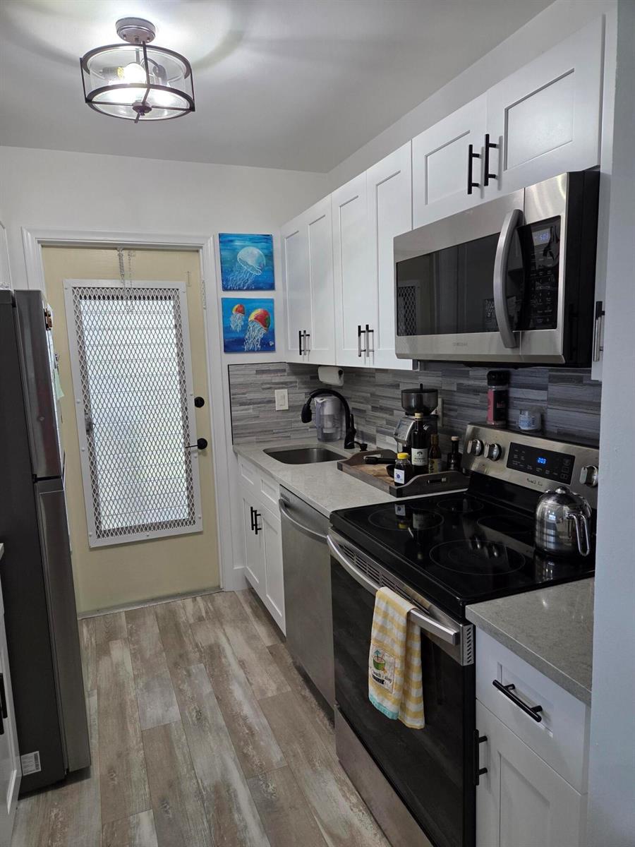 A galley kitchen features white cabinets, stainless steel appliances, a gray backsplash, and wood-look flooring — BOYNTON BEACH, FL