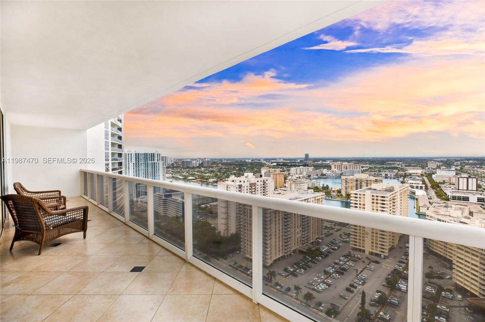 A high-rise balcony with wicker chairs overlooks a city skyline and a waterway at sunset — 1830 S Ocean Dr # 2808, Hallandale Beach FL 33009, HALLANDALE BEACH, FL