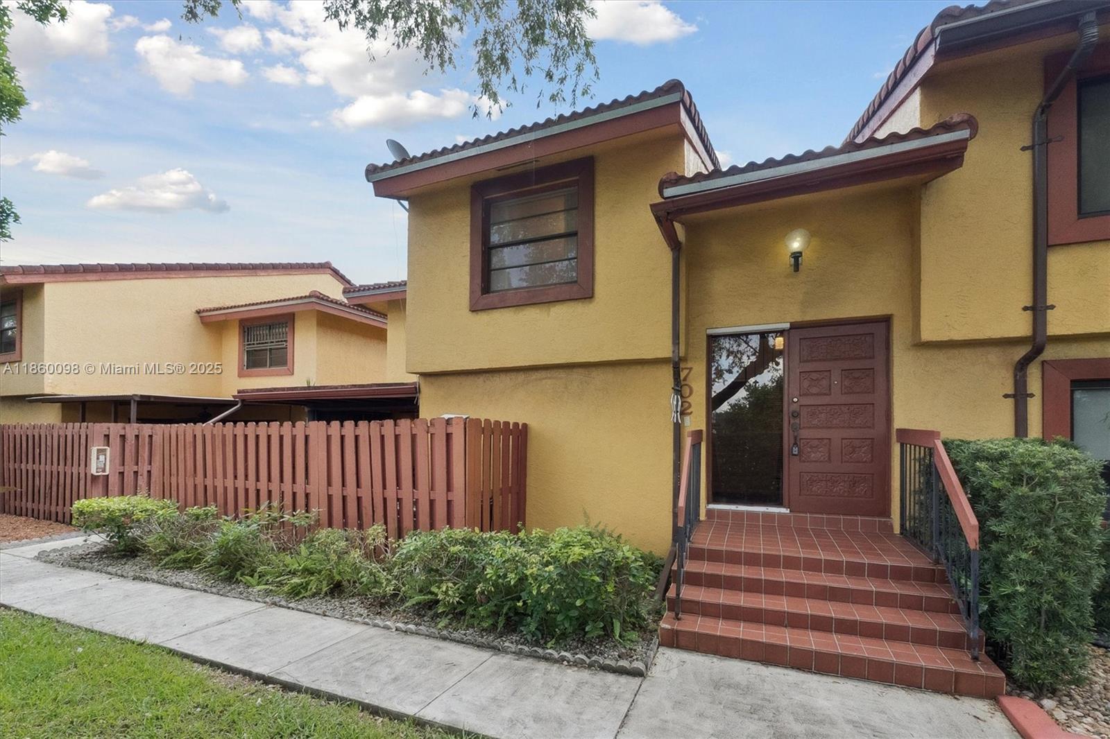 The exterior of a yellow two-story townhouse features a brown wooden fence and tiled steps — 5975 SW 137th Ave # 702, Miami FL 33183, MIAMI, FL