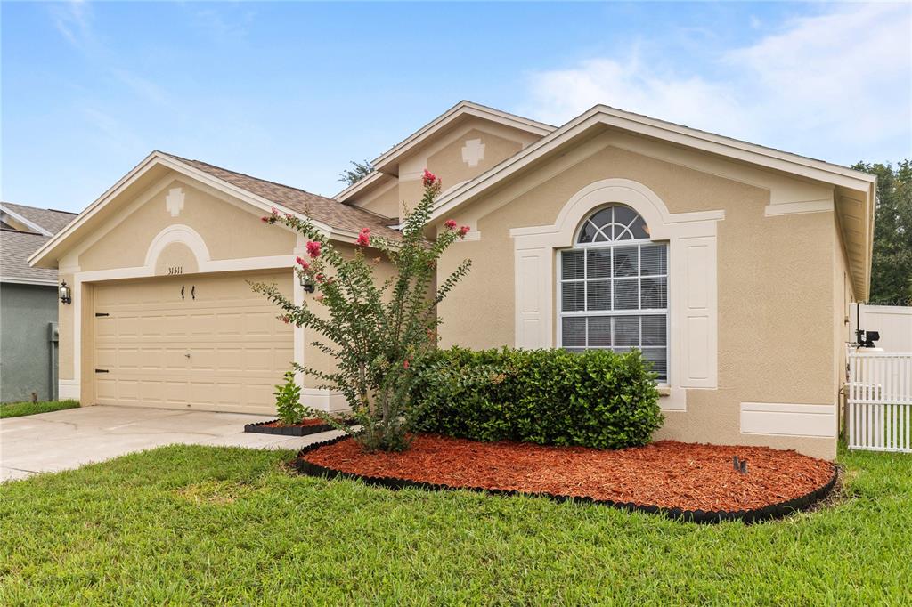 A tan single-story house with a two-car garage, arched window, and a landscaped front yard — 31511 WRENCREST DRIVE, WESLEY CHAPEL, FL