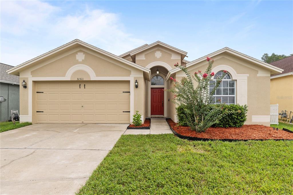 A single-story house with a tan stucco exterior, a red front door, and a two-car garage — 31511 WRENCREST DRIVE, WESLEY CHAPEL, FL
